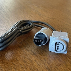 May include: A black electrical power cable featuring a cylindrical grey connector with three circular pin holes and a separate square grey panel-mount socket component that also contains a three-pin configuration for electrical connectivity.