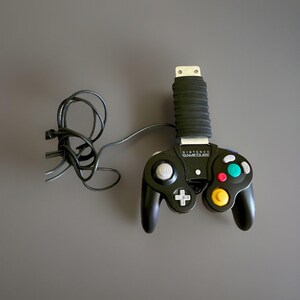 Könnte beinhalten: Ein schwarzer Nintendo GameCube-Videospielcontroller mit einem grauen Analogstick, einem gelben C-Stick, einem weißen Steuerkreuz und bunten Aktionstasten. Ein schwarzes Kabel ist angeschlossen und ein schwarzer gerippter Griff ist oben befestigt.