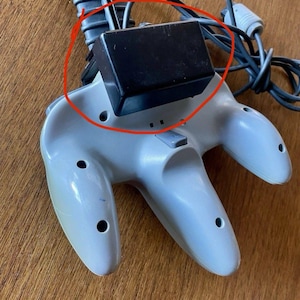 Puede incluir: Un mando de videojuegos gris claro con un paquete de expansión rectangular negro insertado en la ranura trasera. El mando tiene un diseño de tres puntas con orificios para tornillos visibles y un cable gris que sale de la parte superior.