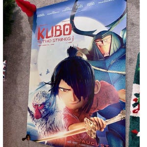 K&ouml;nnte beinhalten: Ein Filmplakat f&uuml;r "Kubo - Der tapfere Samurai" mit einem jungen Mann mit Schwert, einem Affen und einer gro&szlig;en blauen Figur. Das Plakat zeigt den Filmtitel und das Erscheinungsdatum, 19. August.