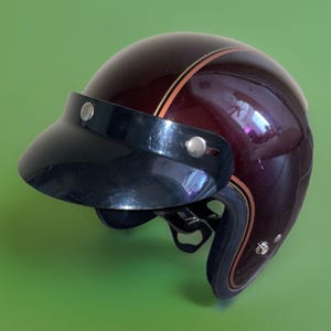 Può includere: Un casco da moto aperto di colore marrone scuro lucido, dotato di una visiera nera fissata con due bottoni a pressione in metallo argentato. Il casco è decorato con sottili strisce parallele color beige e nero che seguono la curva della calotta.