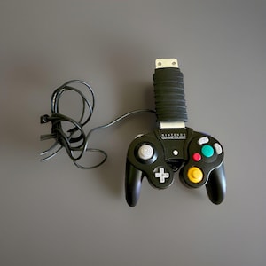 May include: A black Nintendo GameCube video game controller featuring a central black grip attachment, a grey analogue stick, a grey D-pad, a yellow C-stick, a green A button, a red B button, and a black cord extending from the left side of the device.
