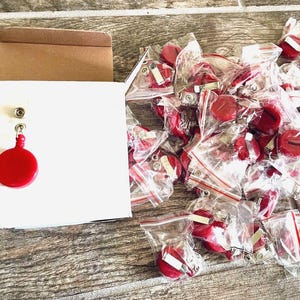 May include: A collection of red, circular objects, possibly buttons or charms, are displayed. Some are individually packaged in clear plastic bags. One is attached to a white card. The items are on a wooden surface.