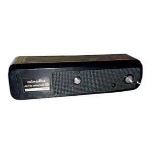 Puede incluir: Un accesorio de cámara negro Minolta Auto Winder G. El dispositivo tiene un agarre texturizado, dos diales plateados y el texto "minolta AUTO WINDER G JAPAN" impreso en una etiqueta. La parte superior es de un material liso y oscuro.