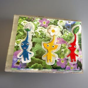 May include: A rectangular clear acrylic block featuring three embroidered patches of Pikmin characters. A blue Pikmin with a white bud, a yellow Pikmin with a white flower, and a red Pikmin with a green leaf are displayed over a floral background.