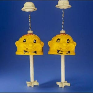 Könnte beinhalten: Zwei gelbe Vintage-Spielzeuge aus Kunststoff mit lächelnden Gesichtern, Fliegen und weißen Stielen. Jedes Spielzeug hat einen weißen Hut, der mit einem gedrehten Metalldraht über dem Kopf aufgehängt ist. Die Spielzeuge stehen vor einem blauen Hintergrund.