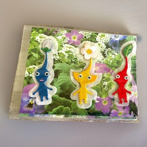 May include: Three embroidered patches featuring blue, yellow, and red Pikmin characters with white flower or leaf sprouts on their heads, displayed on a rectangular card with a printed background of green foliage and purple and white flowers.