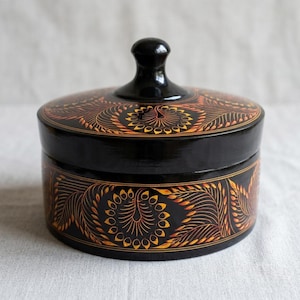 Peut inclure: Un récipient rond noir avec couvercle, orné d'un motif floral peint à la main en orange et jaune. Le couvercle possède un bouton central, et la base cylindrique est décorée de motifs de feuilles répétitifs sur toute la surface extérieure.