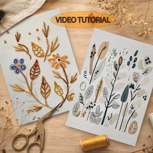 May include: Two rectangular white cards featuring embroidered floral and botanical designs. The left card displays blue and orange flowers with brown stems. The right card shows grey and beige foliage. Includes gold scissors and thread spools.