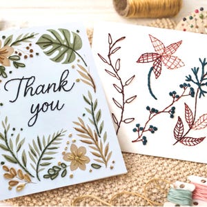 Può includere: Due biglietti di auguri fatti a mano con ricami floreali. Un biglietto dice "Thank you" in corsivo nero, circondato da fiori e foglie ricamati in verde, beige e oro. L'altro biglietto presenta disegni floreali ricamati in rosso, blu e marrone.