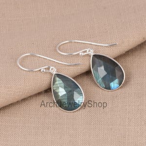 Puede incluir: Un par de pendientes plateados con forma de lágrima y piedras de labradorita facetadas e iridiscentes. Los pendientes se muestran sobre una tela de tono neutro. El texto "Arch Jewelry Shop" es visible.