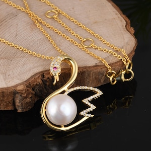 Può includere: Una collana color oro con un ciondolo a forma di cigno. Il cigno è decorato con piccoli cristalli trasparenti lungo il collo e l'ala, una singola pietra rosa per l'occhio e una grande perla bianca rotonda al centro del corpo del cigno.