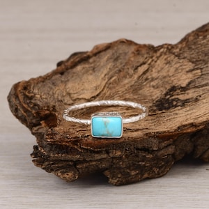 May include: A silver ring with a rectangular turquoise stone. The band has a textured design. The ring is displayed on a piece of wood with a light-colored background. The turquoise stone is a bright, vibrant blue.