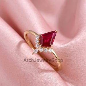 May include: A gold ring featuring a large kite-shaped red gemstone as the centrepiece, accented by a cluster of small round and marquise-cut white stones along one side of the setting. The text ArchJewelryShop is visible on the lower portion.