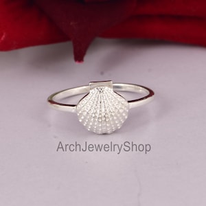 May include: A silver ring featuring a detailed seashell design. The ring has a delicate band and a textured, three-dimensional seashell centerpiece. The ring is displayed on a light surface with a red background.