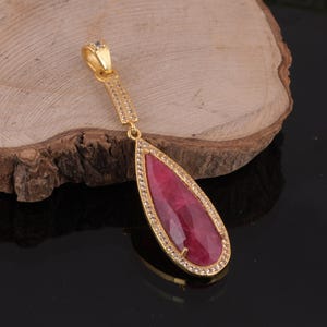 May include: A gold-toned pendant featuring a large, teardrop-shaped, deep pink gemstone set in a frame of small, clear, sparkling stones. The pendant includes a long, slender, rectangular gold-toned bail accented with tiny clear stones.