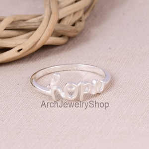 May include: A silver ring with the word "hope" in a cursive font. The ring is on a neutral-colored surface with the text "Arch Jewelry Shop" below the ring. The background includes a woven basket.