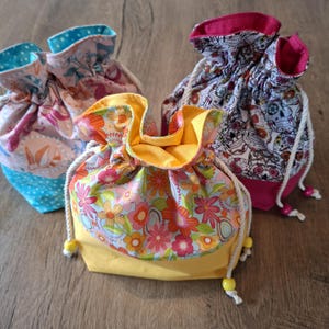 May include: Three fabric drawstring bags in various floral and skull patterns. The bags feature a yellow, pink, and blue colour scheme. Each bag has a drawstring closure with a white cord and yellow beads. The bags are made of fabric.
