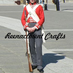 Può includere: Una persona in uniforme militare rossa con dettagli bianchi, pantaloni grigi e scarpe nere, che tiene un fucile. L'uniforme comprende una fascia bianca e un cappello nero alto. Il testo "Reenactment Crafts" è visibile.
