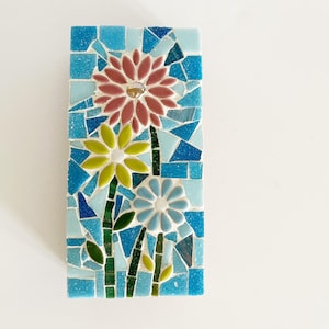 May include: A rectangular mosaic art piece featuring three stylized flowers. The flowers are in shades of pink, yellow, and blue, with green stems and leaves. The background is a mosaic of blue tiles. The artwork is likely for home decor.