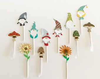 May include: A collection of whimsical garden stakes featuring gnomes, mushrooms, and sunflowers. The gnomes have various hat colors and designs, while the mushrooms and flowers have detailed patterns. Each stake is on a white stick.