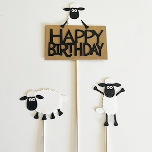 May include: A set of three birthday cake toppers featuring sheep designs. One topper has a brown sign with the words "HAPPY BIRTHDAY" in black. The other two toppers are sheep with white wool and black faces and legs.