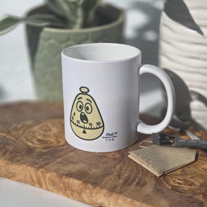 Könnte beinhalten: Weiße Keramik-Tasse mit einem Cartoon-Timer-Design mit überraschtem Gesichtsausdruck. Die Tasse hat einen gebogenen Griff und den Text "Mel's Drama-Laden". Die Tasse steht auf einem Holzbrett mit einem Teebeutel.