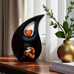 May include: Black teardrop-shaped decorative urn with a heart-shaped accent and a cartoon image of a character in orange and blue. The urn is displayed on a wooden table near a window with a gold vase.