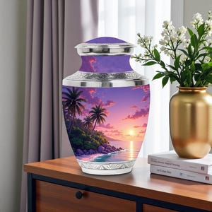 Könnte beinhalten: Dekorative Urne mit einer lebendigen Sonnenuntergangsszene mit Palmen und einem Strand. Die Urne hat einen lila Deckel und Hals, mit silberfarbenen Bändern und einem weißen Sockel. Die Urne wird auf einer Holzoberfläche ausgestellt.