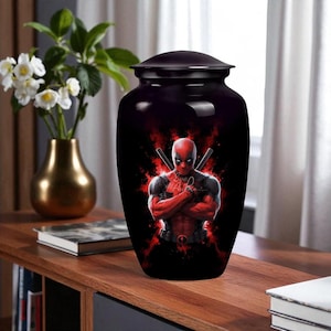 Puede incluir: Urna negra con acabado brillante, con un gráfico de Deadpool. El diseño incluye una combinación de colores rojo y negro con la imagen del personaje y espadas cruzadas. La urna tiene una tapa a juego.