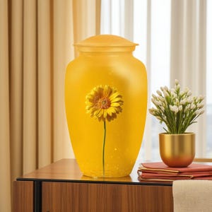 May include: A yellow urn with a lid, featuring a large yellow flower. The urn is on a wooden surface, with a small gold pot of white flowers and a stack of red books nearby. The background includes a window and curtains.