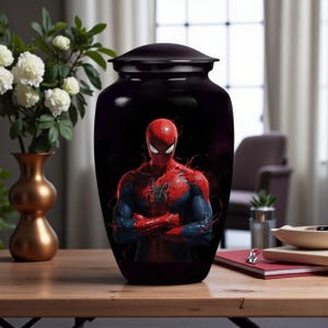Puede incluir: Urna negra brillante con una imagen de Spider-Man con su traje rojo y azul. El superhéroe se muestra con los brazos cruzados sobre un fondo oscuro. La urna está sobre una mesa de madera, con un jarrón de flores blancas en el fondo. La urna tiene tapa.