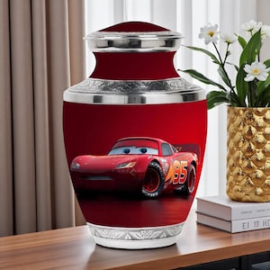 May include: A red urn with a silver rim and lid, featuring a graphic of the animated character Lightning McQueen from the movie Cars. The urn has a glossy finish and is displayed on a wooden surface.