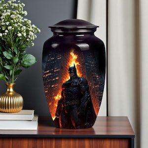 May include: A dark purple urn with a lid, featuring a Batman graphic against a backdrop of a burning city. The superhero is depicted in his iconic black suit, standing amidst flames. The urn is displayed on a wooden surface.