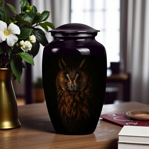 Könnte beinhalten: Schwarze Urne mit Deckel, verziert mit einem detaillierten Eulenporträt. Die Eule hat braune und beige Federn mit stechenden roten Augen. Die Urne steht auf einem Holztisch, mit einer Vase mit weißen Blumen im Hintergrund.