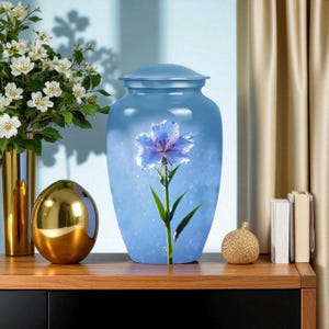 May include: A light blue cremation urn with a lid, featuring a large blue flower design. The urn is displayed on a wooden surface with other decorative items, including a gold vase with white flowers and a gold sphere.