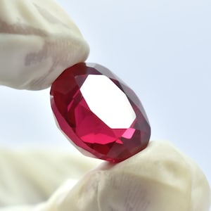 May include: A faceted, oval-shaped ruby gemstone. The ruby is a deep, vibrant red color with multiple facets that reflect light. The gemstone is held between two gloved fingers against a white background.