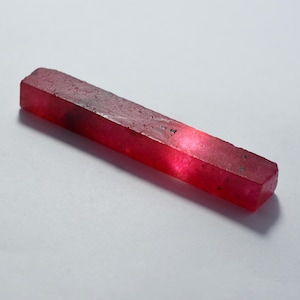 May include: A raw, rectangular ruby gemstone with a deep red hue. The gemstone has a slightly rough, textured surface and appears translucent in the center. The ruby is displayed against a white background.