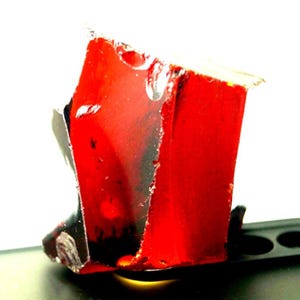 May include: A close-up of a red, rectangular object with a jagged edge. The object appears to be translucent, with dark areas visible inside. The object is resting on a black surface.