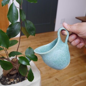 以下が含まれることがあります： 小さな水色のセラミック製じょうろは、観葉植物に水やりをするために使用されています。 じょうろは斑点のある質感と湾曲した注ぎ口を持っています。 植物は緑色の葉を持ち、白い鉢に入っています。