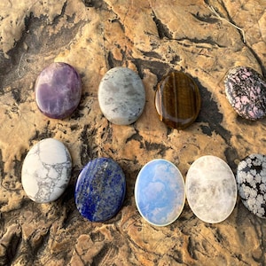Puede incluir: Una variedad de nueve piedras preciosas pulidas en forma ovalada. Las piedras muestran una variedad de colores y patrones, incluyendo púrpura, gris, marrón, rosa, blanco, azul y negro. Las piedras preciosas están dispuestas sobre una superficie texturizada de color marrón claro.