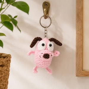 May include: A crochet keychain featuring a pink dog character with large white eyes, black pupils, and dark brown ears and nose. The figure is attached to a silver-toned metal split ring and chain, hanging from a brass-colored wall hook.