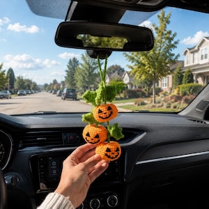 May include: A hanging car accessory featuring three small, orange, crocheted pumpkins with black stitched jack-o'-lantern faces. The pumpkins are connected by a green crocheted vine with several green leaves, suspended from a green yarn loop.