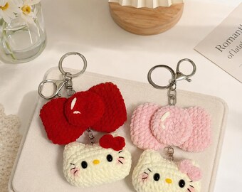 May include: Two crochet keychains featuring white cat faces with black eyes, yellow noses, and pink whiskers. One keychain has a large red bow, and the other has a light pink bow. Both include silver metal key rings and lobster claw clasps.