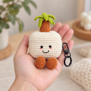 May include: A cream-colored crochet case for wireless earbuds shaped like a palm tree. It features a smiling face with black eyes and pink cheeks, a brown trunk, green palm leaves, and two brown feet. A black metal keychain is attached to the side.