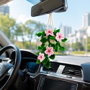 May include: A crocheted hanging plant ornament featuring light pink flowers with dark pink centers and deep green leaves, suspended by a cream-colored cord from a rearview mirror inside a vehicle, showcasing intricate yarn textures and craftsmanship.