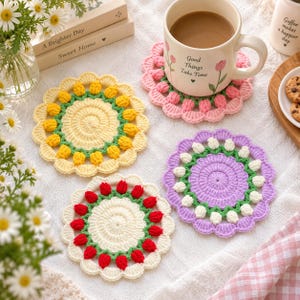 May include: Three round crochet coasters featuring a central circular pattern surrounded by a ring of small, raised flower buds in yellow, red, and white, with scalloped edges in matching colors, arranged near a ceramic mug with tulip graphics.