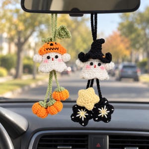 May include: Two crocheted ghost ornaments hanging from a rearview mirror. The left ghost wears an orange pumpkin hat with green vines and dangling orange pumpkins. The right ghost wears a black witch hat with dangling yellow and black stars.