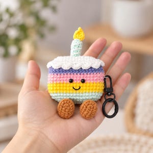 May include: A crochet case for wireless earbuds shaped like a birthday cake with horizontal stripes in pastel purple, pink, yellow, and light green. It features a white scalloped top, a yellow candle, two brown feet, and a black metal keychain.