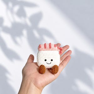 May include: A small, handmade crochet plush toy shaped like a piece of sushi. The body is white with a smiling face featuring two black bead eyes and a stitched mouth. It has a pink textured top and two small brown feet at the bottom.